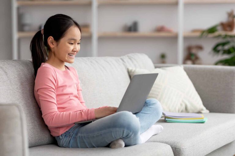 Een kind die blij een website aan het ontwerpen is op een grijze bank met een blauwe broek en roze shirtje aan.